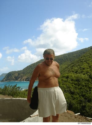 This is a personal photograph showing a middle-aged man shirtless at what appears to be a tropical or Mediterranean coastal location with dense green vegetation and turquoise water visible in the background. The image appears to be a casual vacation 