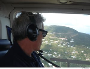 This is an aerial photograph taken from inside a helicopter, showing a person wearing headphones and sunglasses looking out at a mountainous landscape dotted with residential properties. The image appears to be documentation of aerial surveillance or
