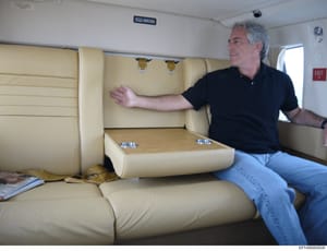 This photograph shows the interior of a private aircraft cabin, featuring cream-colored leather seating and a table surface. A middle-aged man with gray hair, wearing a black polo shirt and jeans, is seated in the cabin gesturing with his hand. The i