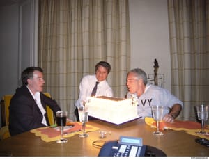 A photograph of three men in a formal meeting setting, seated at a table with glasses of water and what appears to be official documents. The setting suggests an official or business meeting, possibly a deposition, interview, or formal discussion. A 