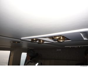 This photograph shows the interior ceiling of an aircraft, featuring recessed brass or gold-toned lighting fixtures embedded in the white ceiling panel with trim. The image captures the cabin interior during what appears to be daytime flight, with a 