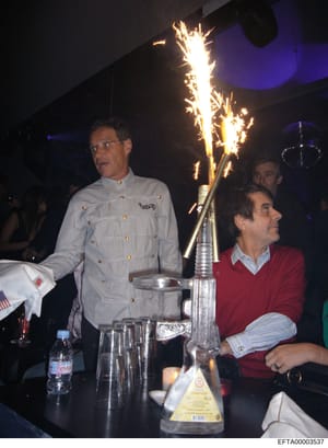 This photograph shows an indoor social gathering or party with at least three visible attendees around a table with a dramatic flaming centerpiece. The man on the left wears a distinctive light blue military-style jacket with gold buttons, while othe