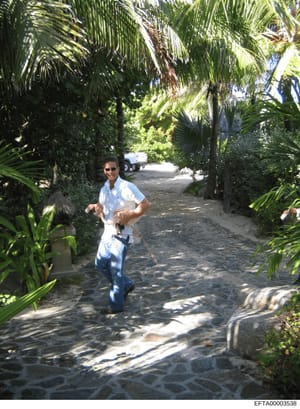 A photograph showing an unidentified man jogging on a stone driveway lined with tall palm trees and dense tropical vegetation. A vehicle is visible in the background. This appears to be evidence photo documentation of a residential property, likely o