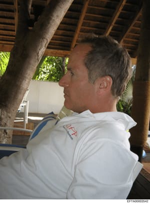 This is a photograph showing a middle-aged man wearing a white polo shirt with a red and blue logo/emblem, photographed in profile at what appears to be a covered outdoor structure or pavilion with wooden beams and tropical vegetation visible. The im