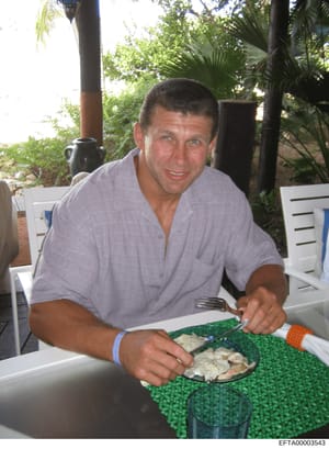 This is a personal photograph showing a man dining outdoors in a tropical setting with palm trees and lush vegetation visible in the background. The photo appears to be a casual social gathering or meal, captured in daylight. The image is identified 