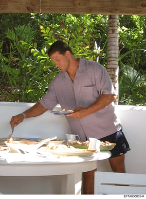 This photograph shows a male staff member in a light purple polo shirt serving or preparing food at an outdoor dining area with a white table. The setting appears to be a tropical property with lush vegetation, palm trees, and a brick column visible 