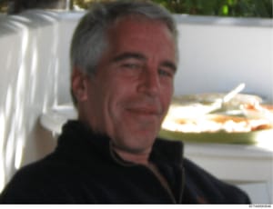 This is a photograph showing a middle-aged man with gray hair wearing dark clothing, photographed outdoors near what appears to be a catered event or gathering. A white structure is visible in the background along with plates of food. The image appea