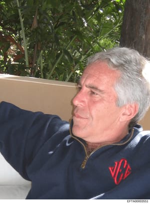 This is a candid photograph of an older male individual wearing a dark blue zip-up jacket with a red logo, seated outdoors against a backdrop of tropical vegetation and a light-colored wall. The image appears to be a personal or documentary photograp