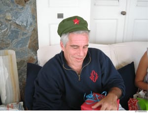 This photograph shows an older man seated indoors wearing a green military-style cap with a red star and a dark blue jacket with red monogrammed initials. The image appears to be from a casual indoor setting with white walls, doors, and decorative st