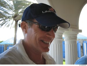 This is a casual photograph showing a middle-aged man wearing a dark blue cap with 'LIMITI' branding and dark sunglasses, smiling at an outdoor location. The setting appears to be a tropical waterfront property with palm trees, white classical column