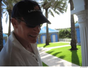 This is a photograph showing a man wearing a dark cap with 'LIMITLESS' text and a white shirt, standing in what appears to be the courtyard or grounds of a luxury Palm Beach residence. The image shows manicured lawns, palm trees, and distinctive blue