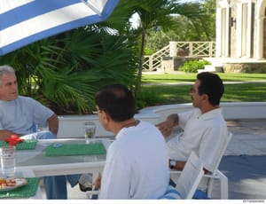 This photograph shows three men seated at a white table under a blue and white striped awning at what appears to be a luxury tropical property. The setting includes palm trees, manicured lawns, and a large mansion or estate building visible in the ba