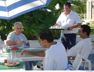 This photograph shows four men at an outdoor gathering under a blue and white striped umbrella at what appears to be a tropical residence. The setting includes a table with refreshments and snacks, palm trees in the background, and manicured grounds.