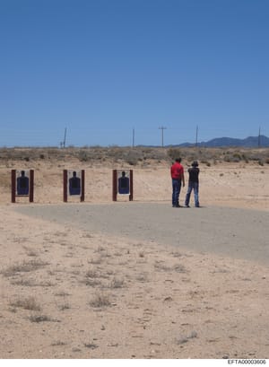This is a photograph of an outdoor shooting range in a desert landscape. The image shows three wooden target frames with blue silhouette targets, and two individuals observing from a distance. The location appears to be a remote, arid area with spars