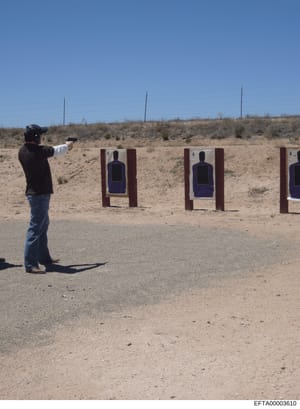 This photograph shows a person engaged in firearms training at an outdoor desert shooting range. The image depicts proper shooting stance and form at what appears to be a professional or law enforcement training facility, with multiple target station