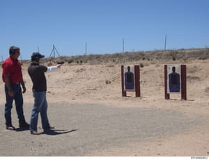 This photograph shows two men at an outdoor shooting range in an arid, desert-like environment. One man in a red shirt observes while another in a black shirt and cap aims a handgun at two target silhouettes mounted on wooden frames. The image appear