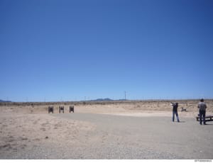 This is a photograph of an outdoor desert range facility, likely used for firearms training or testing. The image shows three target frames set up in an arid landscape with two individuals visible in the scene. The location appears to be a remote, sp