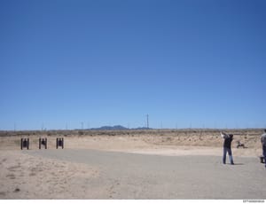 This is a photograph of an outdoor shooting range located in an arid, desert environment. The image shows three black target stands set up on a concrete pad, with a person in dark clothing positioned at a firing line. The landscape consists of sparse
