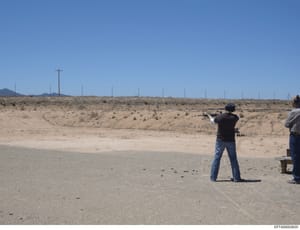 This photograph documents an outdoor shooting range in a desert landscape, showing an individual firing a rifle at targets. The image captures range safety practices and appears to be documentation of firearms activity, possibly related to investigat