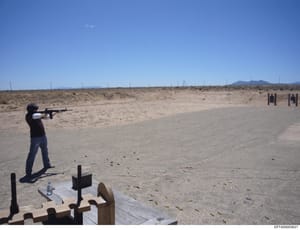 This photograph documents an individual at an outdoor desert shooting range, firing a rifle at target stations visible in the distance. The image shows spent ammunition casings on the ground and appears to be from a firearms training or practice faci