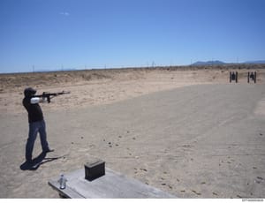 This photograph documents a person engaged in target shooting at an outdoor desert shooting range. The image shows a shooter in firing stance with an AR-style rifle aimed at distant targets. The scene includes spent ammunition casings on the ground, 