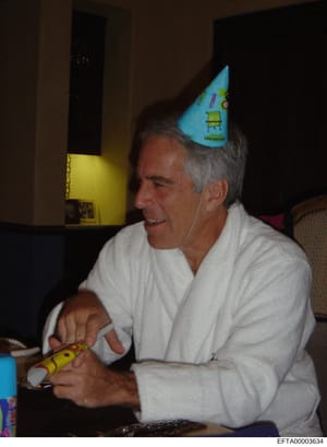 This is a candid photograph showing a middle-aged man wearing a blue party hat at what appears to be an indoor celebration or gathering. The man is seated, wearing a white long-sleeved shirt, and holding what appears to be a toy or novelty item. The 