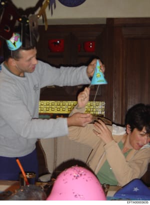 This photograph shows an indoor social gathering or party, likely taking place in a residential setting. An adult male in a gray shirt is interacting with others at a table decorated with party supplies. The image captures what appears to be a casual