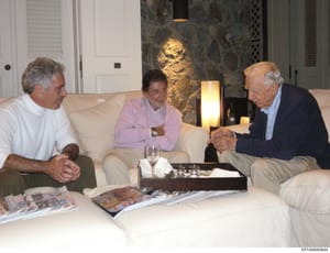 This photograph shows three men in casual conversation seated on cream-colored sofas in a luxuriously appointed interior space. The setting features contemporary artwork, accent lighting, and high-end furnishings consistent with an affluent residenti