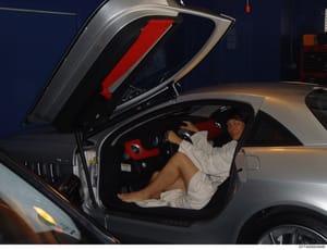 This is a photograph showing an individual posing inside a silver sports car with distinctive red and black interior trim. The vehicle door is open, revealing custom red seat upholstery and black accents. The photo appears to be taken in an indoor fa