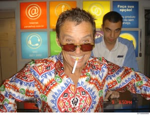This is a casual photograph taken at what appears to be a Brazilian internet café or service business, showing two men posing for the camera. The man in the foreground wears a distinctive colorful patterned shirt and red-tinted sunglasses with a ciga