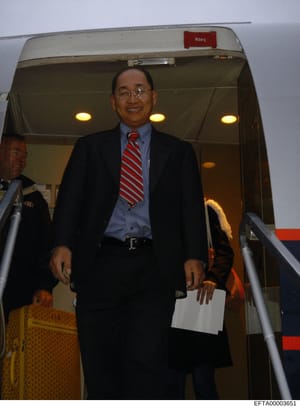 This photograph shows an unidentified Asian male in business attire exiting an aircraft via the stairs. The man wears a dark blazer, light blue dress shirt, and red and white striped tie, and carries what appears to be a white document or folder. The