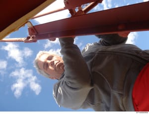 This is a candid photograph showing an older man with white hair on what appears to be a residential property, photographed from below against a blue sky. The image captures an individual near wooden and metal structural elements, possibly a deck or 