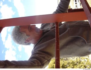 This is a candid photograph showing a man with gray hair wearing dark clothing, positioned near a red metal beam or structure outdoors. The image appears to be taken from a low angle with clear sky visible in the background. The context and relevance