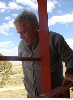 This is a personal photograph showing a middle-aged man with gray hair wearing a gray/dark zippered jacket at what appears to be a rural property. He is positioned next to a large red metal structure (possibly a gate or fence post) with arid grasslan