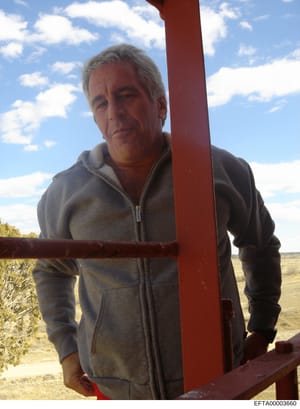 This is a candid photograph showing a middle-aged man with gray hair wearing a gray zip-up hoodie, standing outdoors against a red metal structure with an arid landscape in the background. The image appears to be a personal or surveillance photograph