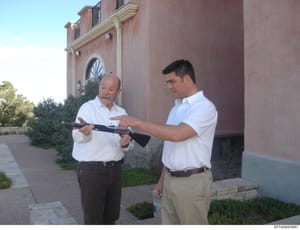 Photograph showing two men in white shirts examining a rifle outside a brick residential building. The image appears to be evidence documentation, possibly related to property inspection or weapons examination. The Bates number indicates this is part
