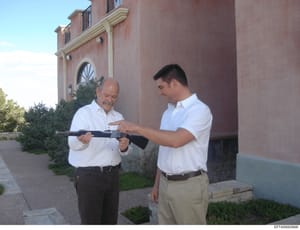 Photograph showing two men in white shirts examining what appears to be an AR-15 style rifle outside a large residential building with distinctive pink/mauve colored brick architecture. The image appears to be documentation of a property or weapon-re
