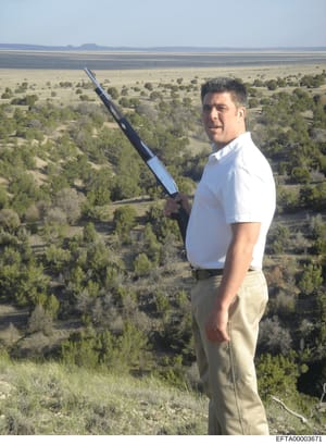 This is a personal photograph depicting an unidentified man standing on an elevated desert terrain, holding what appears to be a sword or large blade. The image shows a vast, sparsely vegetated landscape with scattered trees and mountains visible in 