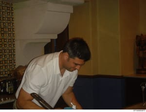 This is a candid photograph showing a male individual in a white polo shirt seated in what appears to be a residential interior. The image captures interior details including patterned wallpaper, furniture, and household items. The photograph's prese