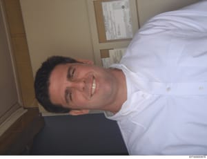 This is a casual personal photograph showing a man in a white shirt smiling at the camera in what appears to be an office or institutional setting. The image is rotated 90 degrees and shows the subject from roughly chest-up. Documents or papers are v