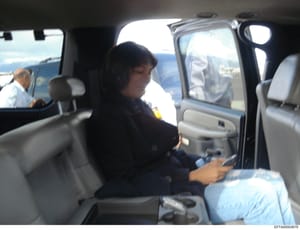 This is a surveillance or investigative photograph showing a person seated in the back seat of a vehicle. The image captures what appears to be monitoring or documentation of movement, with multiple vehicles visible in the background. The photo quali