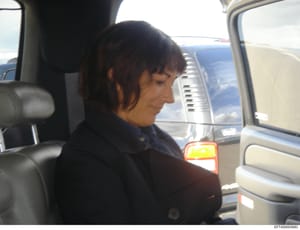 This is a photograph showing a woman with dark hair in profile, seated in the back seat of a vehicle. The image appears to be taken from inside a car, with visible windows, door panels, and another vehicle visible in the background. The photo quality