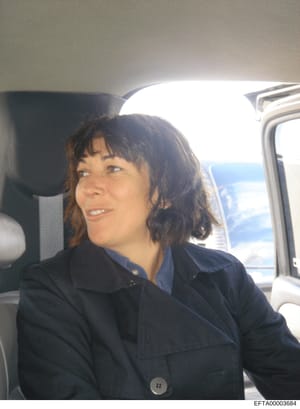 This is a candid photograph of a woman with dark hair with bangs, wearing a dark navy blazer, seated in the passenger seat of a vehicle. The photo appears to be taken during daytime with natural sunlight visible through the windows. The context and s