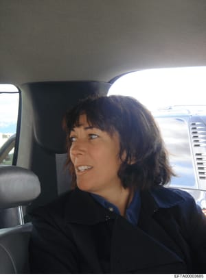 This is a casual photograph of a woman seated in the passenger or rear seat of a vehicle. The image appears to be a personal photo taken during daytime, showing the subject in profile wearing dark clothing. Without additional context or identifying i