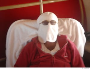 This is an evidence photograph showing a person whose face is deliberately concealed with white cloth wrapping and dark sunglasses. The person wears a red jacket with white sleeves and is seated against a red upholstered background. The image appears
