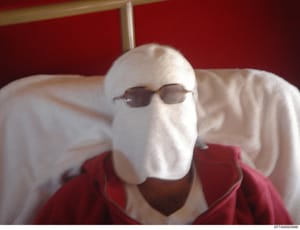This is a photograph showing a person whose face is completely obscured by white cloth wrapping, with only dark sunglasses visible. The person is wearing a red/maroon colored garment and is seated against a red background with white bedding or cushio