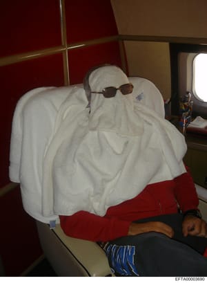 This photograph shows a person seated in what appears to be the interior of a private aircraft, wrapped extensively in white towels with their head covered, wearing dark sunglasses. The setting includes burgundy and tan interior paneling typical of a