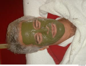 This is a casual personal photograph showing a middle-aged man with gray hair receiving what appears to be a spa facial treatment, with a green face mask applied. The photo is set against a red background with white towels visible. The image's presen
