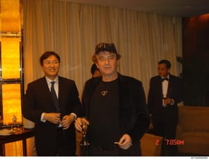 This is a photograph documenting three men at what appears to be a formal indoor event or social gathering. The image shows an elegant interior setting with floor-to-ceiling curtains and illuminated display shelving. The timestamp indicates the photo