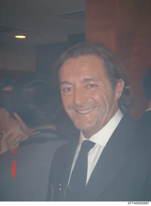 This is a photograph of a middle-aged man wearing a dark suit and blue tie at what appears to be an indoor social or formal event. The image quality is somewhat grainy and the lighting is dim, suggesting it was taken at an evening venue. The photo is